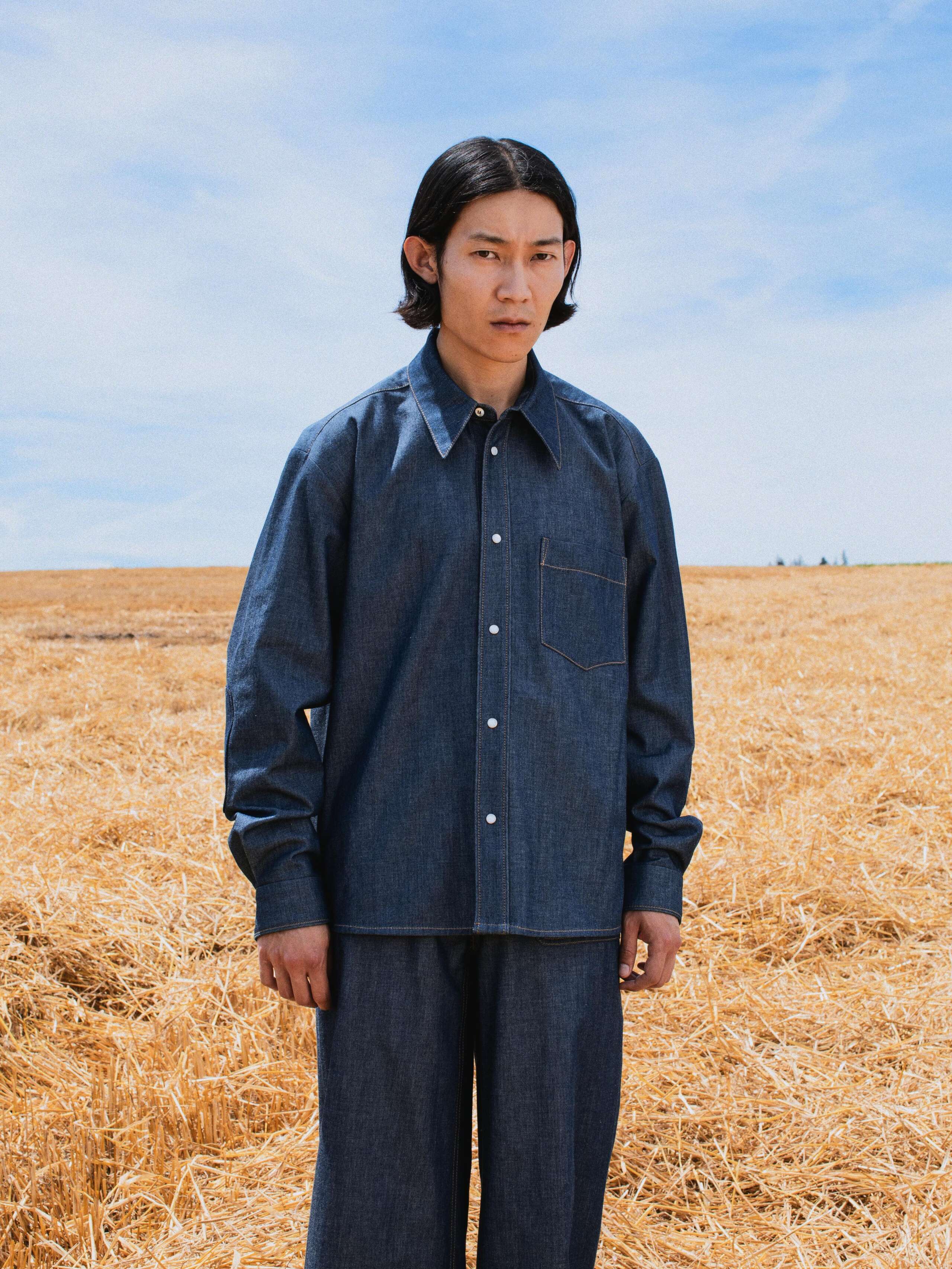 Picture of a male model standing in a yellow corn field wearing a denim shirt and jeans