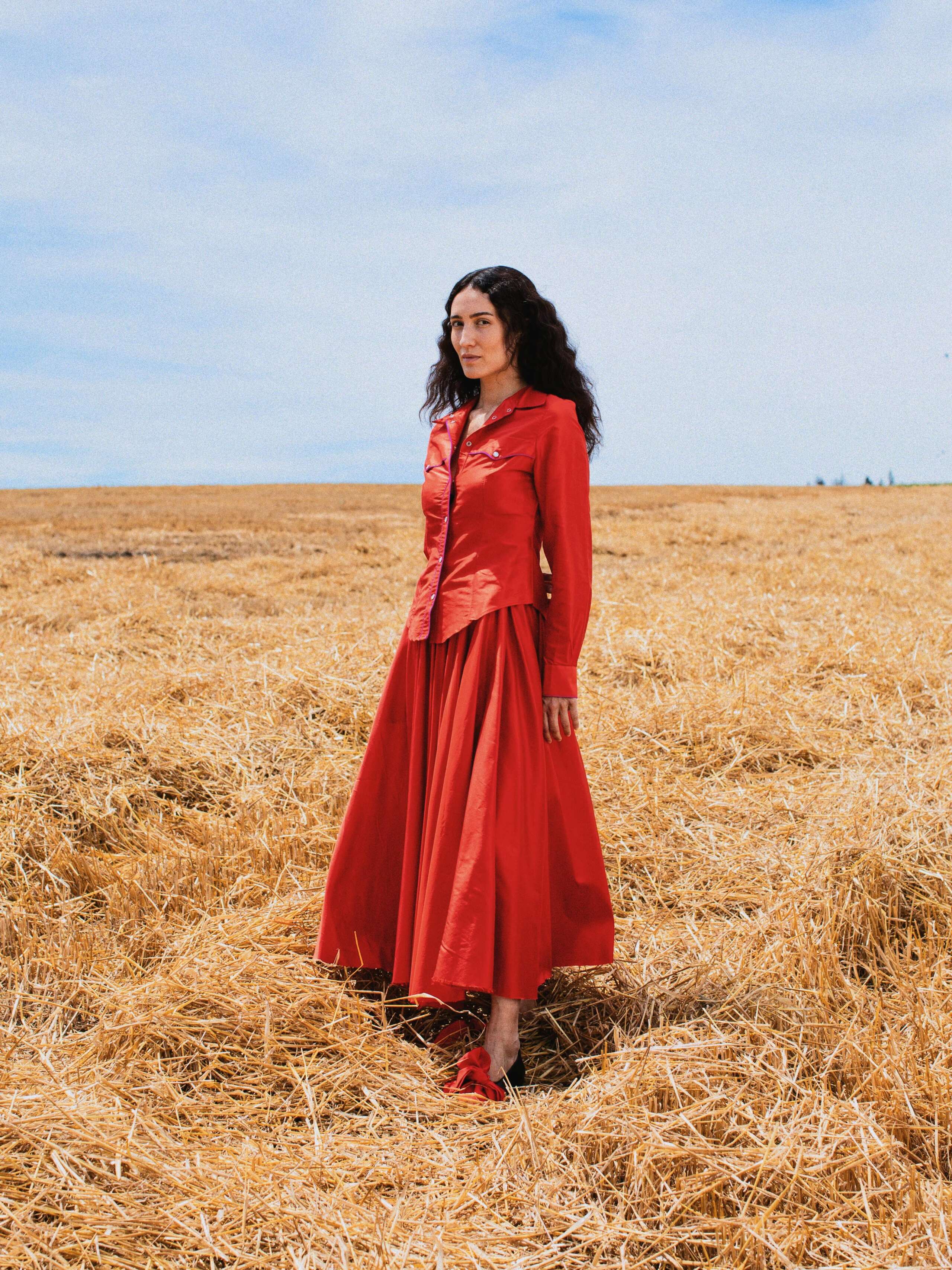 Picture of a female model standing in a yellow corn field wearing a combination of a red silk skirt and a red silk blouse