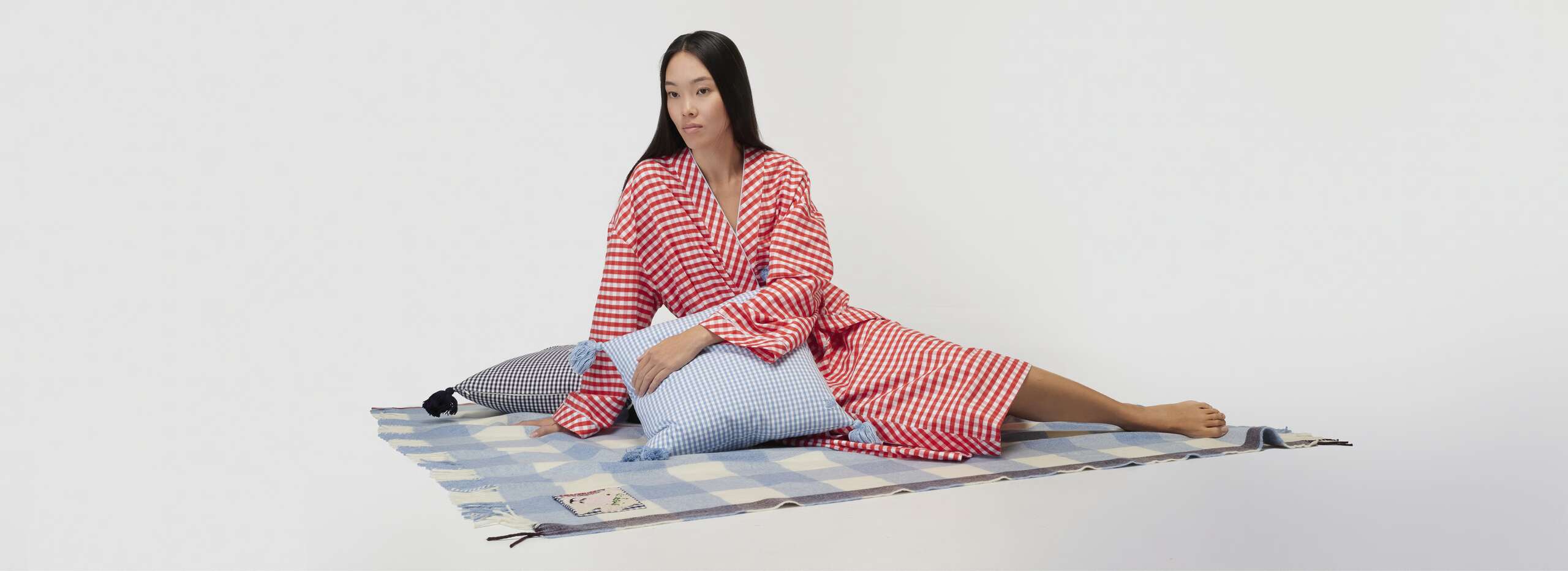 Picture of a women sitting on an enSoie wool blanket, holding a pillow in her arm and wearing a bathrobe featuring a red Vichy check pattern